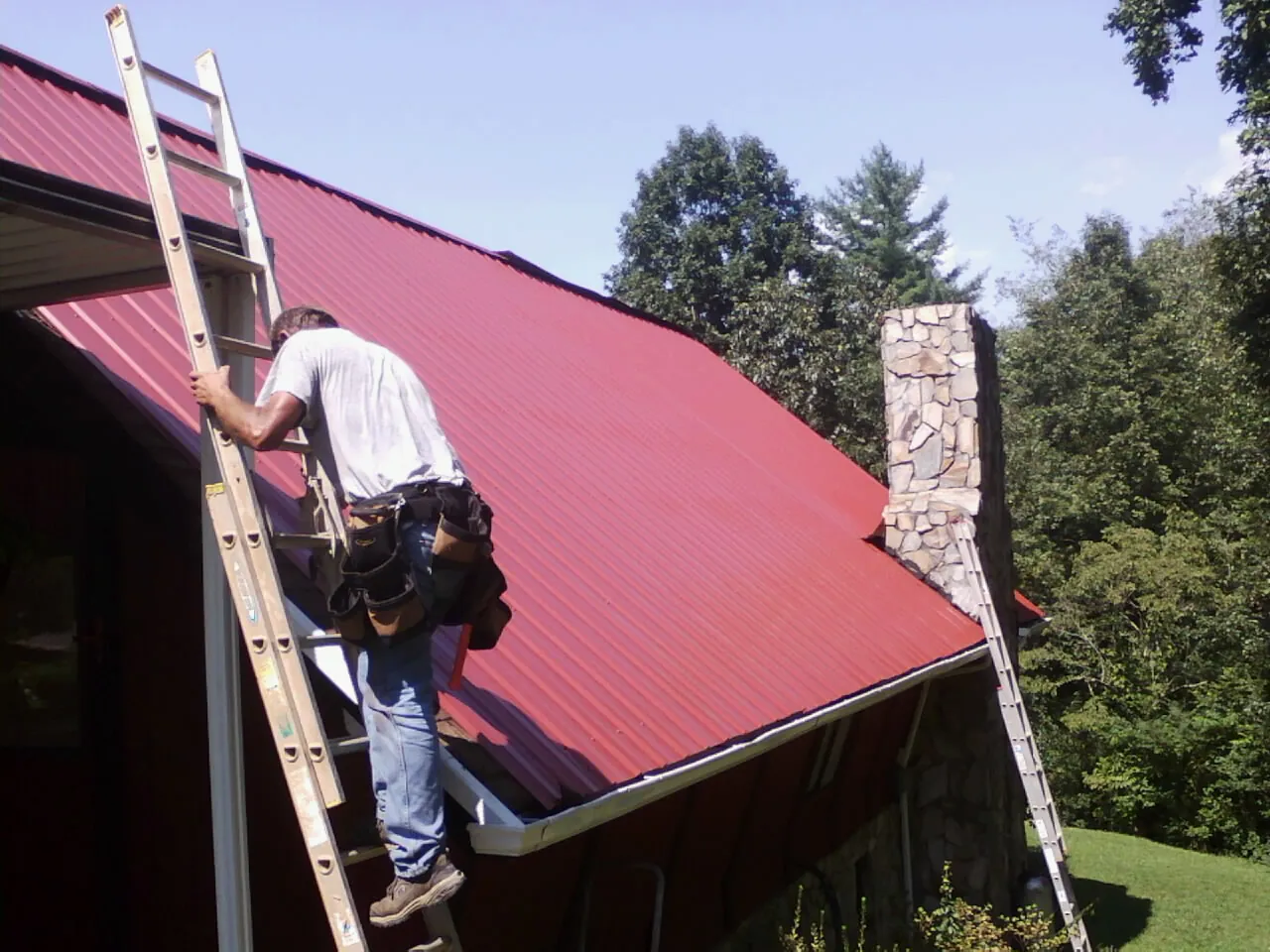 Roofer on a ladder beside a red metal roof and stone chimney — Soffit & Fascia Repair by Roofing Brothers in Castro Valley